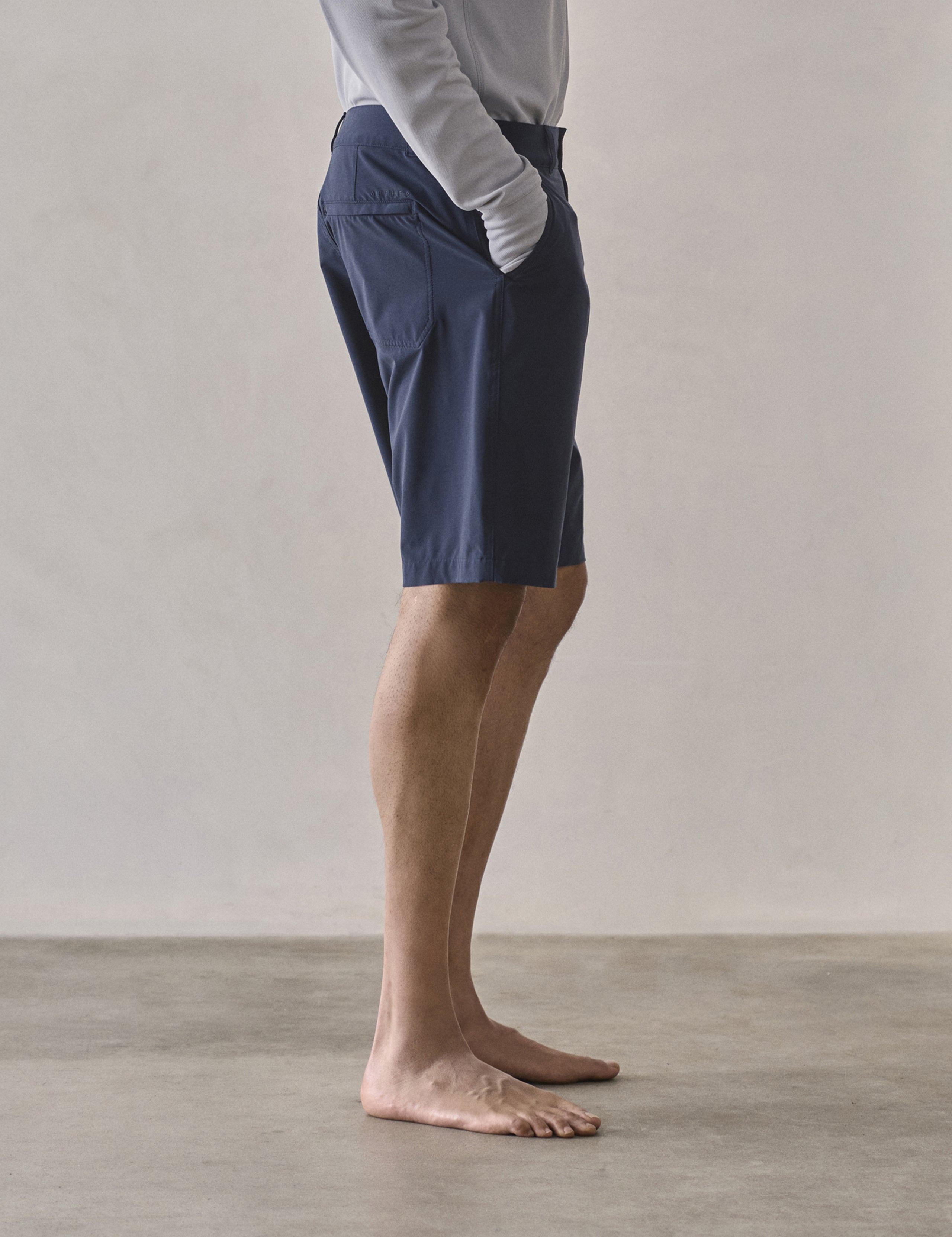Right side profile lower-body view of man with hands in pockets wearing Catalina Submersible Short in Total Eclipse dark blue from AETHER Apparel.