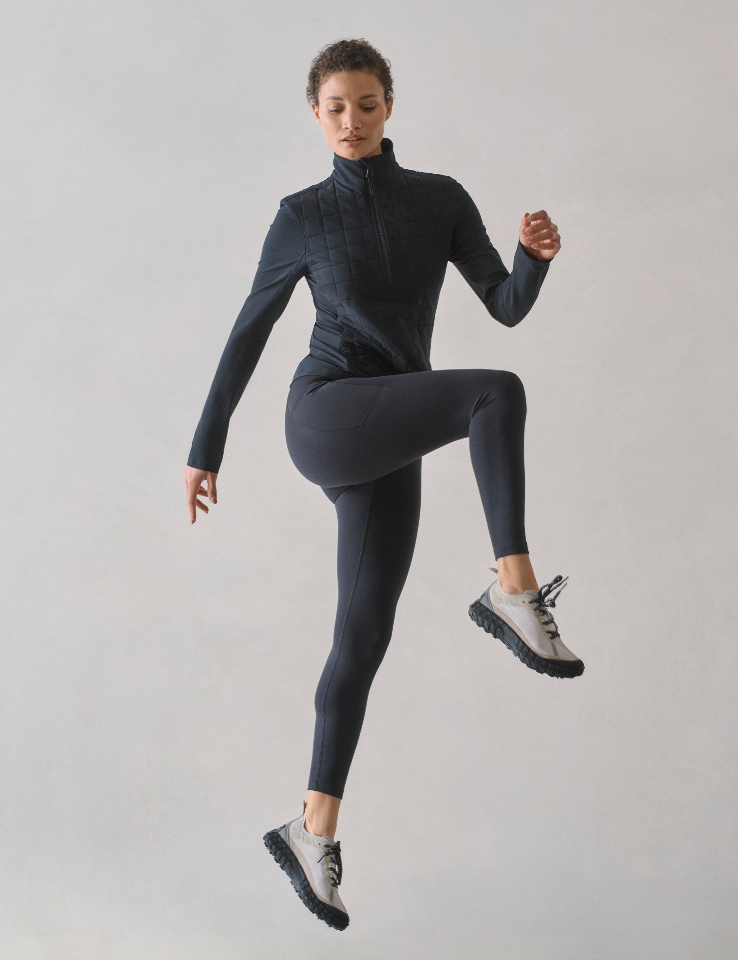 Studio image of woman jumping while wearing Wren Insulated Half-Zip in Onyx Black from AETHER Apparel.